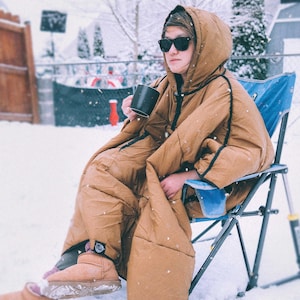 May include: A person wearing a brown hooded sleeping bag suit, black sunglasses, and tan boots, sitting in a blue camping chair in a snowy backyard. They are holding a black mug. The suit has black trim.