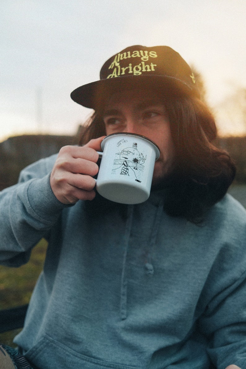 May include: A person wearing a black baseball cap with the text "Always Alright" in yellow embroidery, is drinking from a white enamel mug with a black and white illustration of a person surfing. The person is wearing a light blue hoodie.