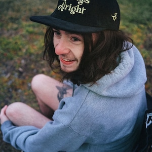 May include: A person wearing a black baseball cap with the text "Always Alright" in yellow embroidery. They are wearing a gray hoodie and sitting on a black chair.