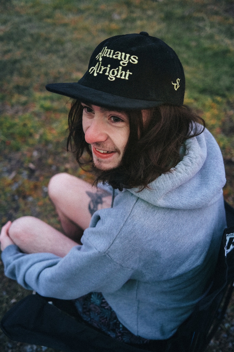 May include: A person wearing a black baseball cap with the text "Always Alright" in yellow embroidery. They are wearing a gray hoodie and sitting on a black chair.