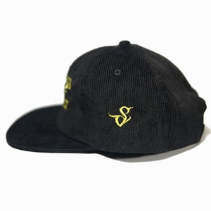 May include: A black corduroy baseball cap with a gold embroidered logo on the side. The logo is a stylized letter "S".