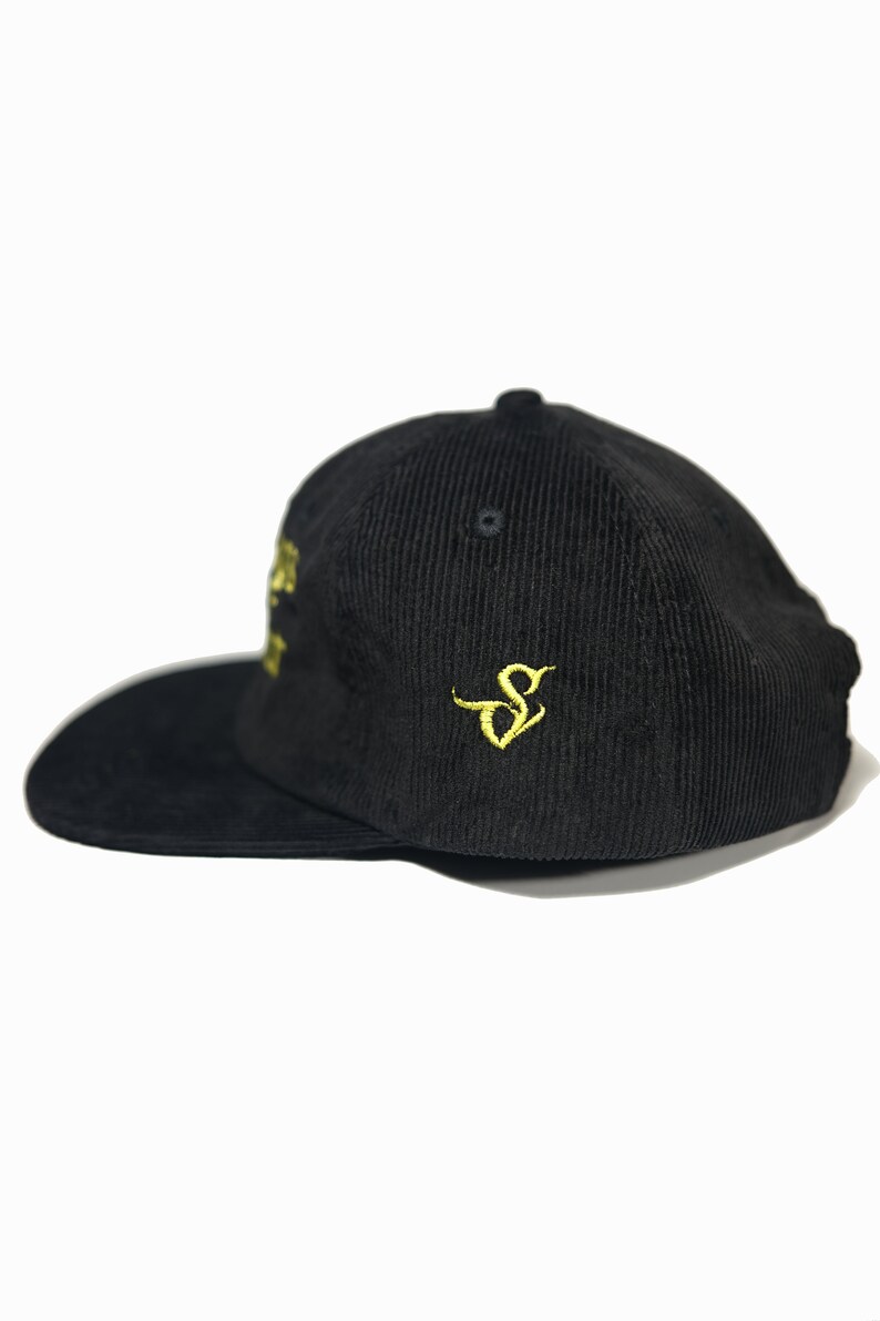 May include: A black corduroy baseball cap with a gold embroidered logo on the side. The logo is a stylized letter "S".
