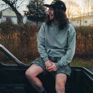 May include: A person wearing a gray hoodie with a black baseball cap that says "Always Right" in yellow letters. They are sitting in the back of a truck and wearing black and white patterned shorts and black and yellow sneakers.