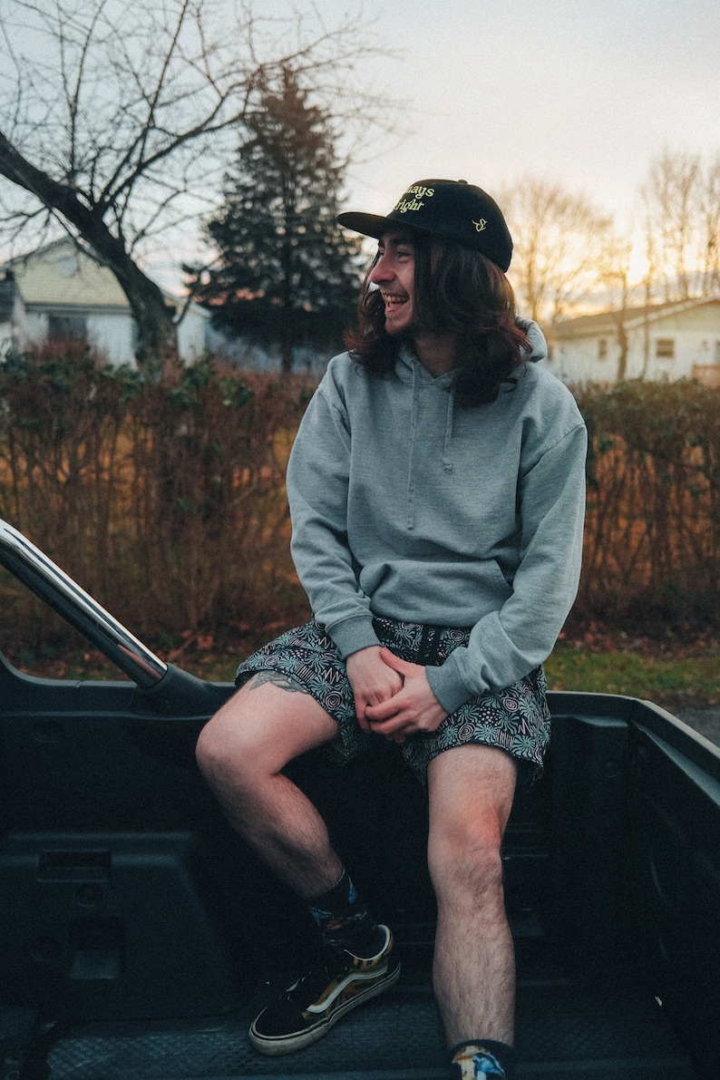 May include: A person wearing a gray hoodie with a black baseball cap that says "Always Right" in yellow letters. They are sitting in the back of a truck and wearing black and white patterned shorts and black and yellow sneakers.