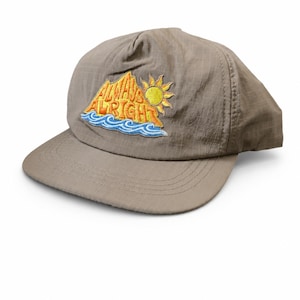 Peut inclure: Une casquette de baseball taupe avec une texture de tissu à motif quadrillé. L'avant présente un écusson brodé représentant un soleil jaune, des montagnes orange et des vagues bleues avec le texte ALWAYS ALRIGHT en lettres orange.