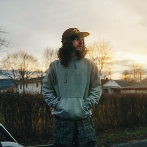 May include: A person wearing a gray hoodie with the text "Mind Over Matter" on a black baseball cap, standing in the back of a pickup truck. The person is wearing black and green patterned shorts.