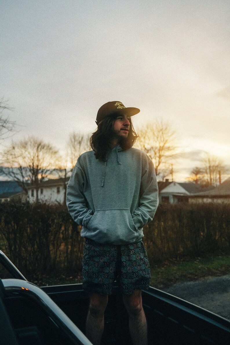 May include: A person wearing a gray hoodie with the text "Mind Over Matter" on a black baseball cap, standing in the back of a pickup truck. The person is wearing black and green patterned shorts.