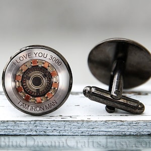 May include: A pair of silver cufflinks with a round, black and copper design. The design features a circular pattern with the text "I love you 3000 I am Iron Man".