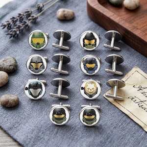 May include: Silver cufflinks featuring various helmet designs in green, blue, white, and gold. The cufflinks are displayed on a grey fabric, with small stones and lavender sprigs in the background.