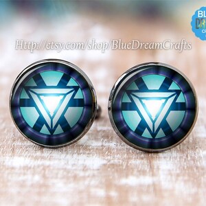 Może przedstawiać: Para mankietów z wzorem niebieskiego i białego reaktora łuku Iron Mana. Spinki do mankietów są srebrne z okrągłym szklanym kaboszonem.