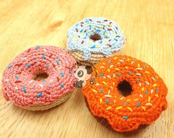 Puede incluir: Tres juguetes de rosquilla tejidos a ganchillo con bases color canela y glaseado rosa, azul claro y naranja. Cada rosquilla está decorada con pequeñas puntadas de hilo multicolor que representan chispas. Se ve un logo con un panda.