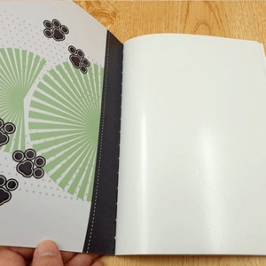 May include: A white notebook with a black spine and a green and black patterned cover. The cover features a design of a fan with black paw prints.