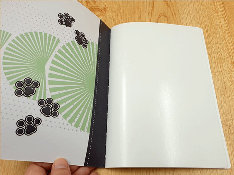 May include: A white notebook with a black spine and a green and black patterned cover. The cover features a design of a fan with black paw prints.