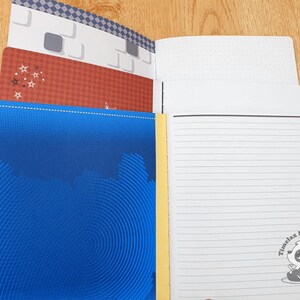 May include: A blue notebook with a yellow spine and a spiral binding. The cover has a pattern of concentric circles. The notebook is open to reveal lined pages with a panda bear logo that says "Timelax Mini Creations".