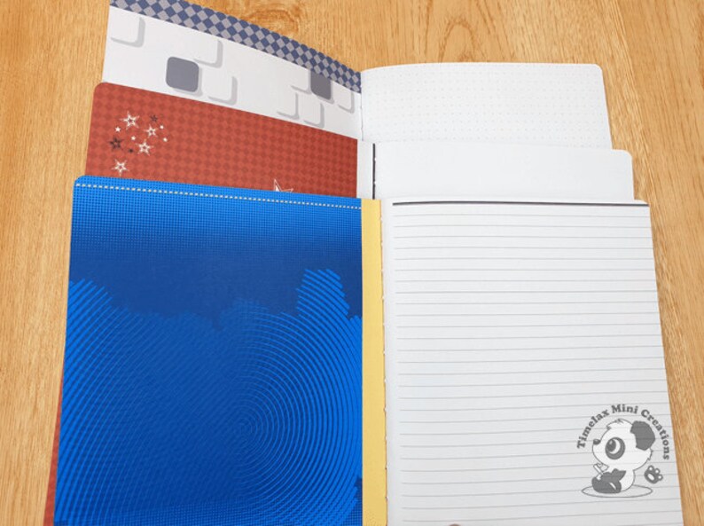 May include: A blue notebook with a yellow spine and a spiral binding. The cover has a pattern of concentric circles. The notebook is open to reveal lined pages with a panda bear logo that says "Timelax Mini Creations".