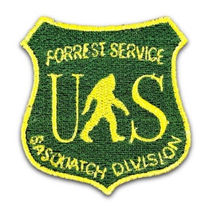 May include: Embroidered patch with a green shield design and yellow border. The patch features the text "FORREST SERVICE" above a yellow silhouette of a Sasquatch, with the letters "UAS" and "SASQUATCH DIVISION" below.