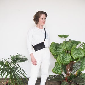 Pode incluir: Uma mulher vestindo uma camisa branca de manga comprida e calças brancas com uma bolsa transversal preta. Ela está em pé em frente a uma parede branca com plantas em vasos.