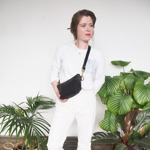 Può includere: Una donna indossa pantaloni bianchi e una camicia bianca a maniche lunghe con una borsa a tracolla nera. È in piedi davanti a un muro bianco con due piante in vaso.