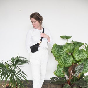 Op de afbeelding: Een vrouw draagt een wit overhemd met lange mouwen en een witte broek met een zwart leren heuptasje dat als schoudertas wordt gedragen. Ze staat voor een witte muur met twee planten in potten.