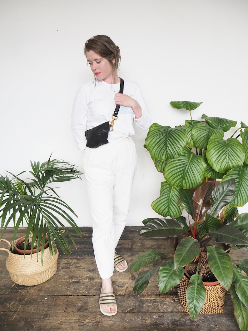 Op de afbeelding: Een vrouw draagt een wit overhemd met lange mouwen en een witte broek met een zwart leren heuptasje dat als schoudertas wordt gedragen. Ze staat voor een witte muur met twee planten in potten.