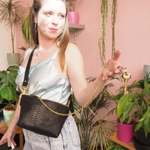 May include: A woman wearing a light blue satin top and denim jeans is holding a black snakeskin crossbody bag with a gold chain strap. The bag is worn on her waist and has a gold zipper. She is holding a set of keys with a gold chain attached to the bag.