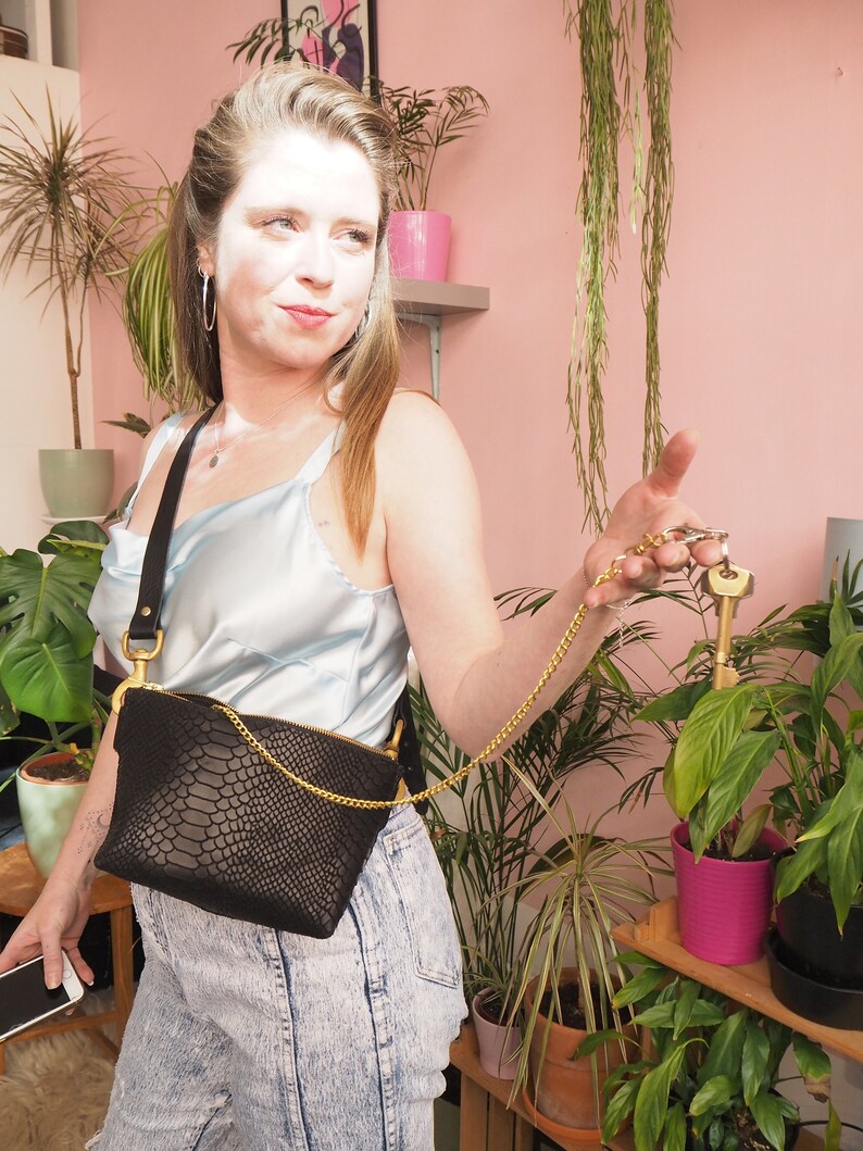 May include: A woman wearing a light blue satin top and denim jeans is holding a black snakeskin crossbody bag with a gold chain strap. The bag is worn on her waist and has a gold zipper. She is holding a set of keys with a gold chain attached to the bag.