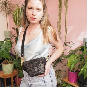 Op de afbeelding: Een vrouw draagt een lichtblauw satijnen topje en een spijkerbroek en houdt een zwarte slangleren schoudertas vast met een gouden ketting en sluiting.