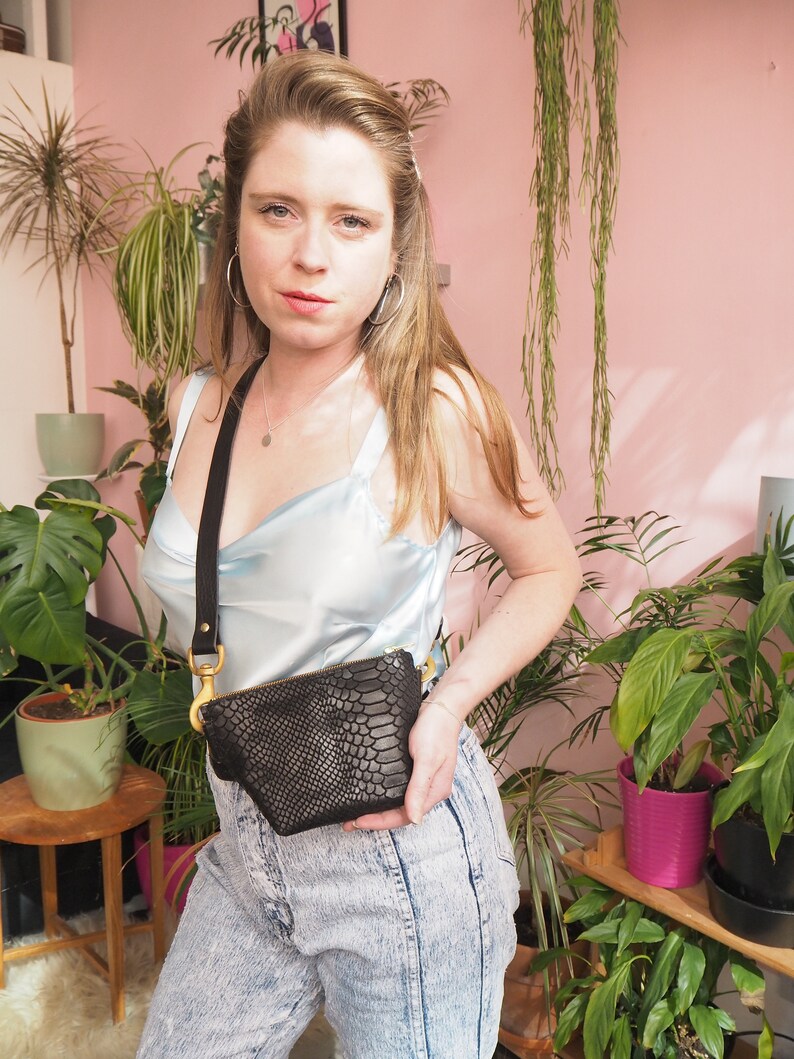 Op de afbeelding: Een vrouw draagt een lichtblauw satijnen topje en een spijkerbroek en houdt een zwarte slangleren schoudertas vast met een gouden ketting en sluiting.