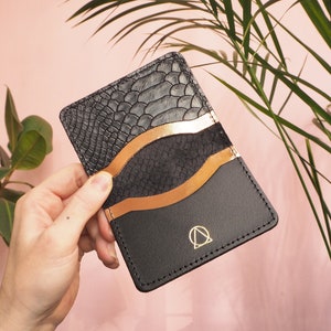 K&ouml;nnte beinhalten: Ein schwarzer Lederkartenhalter mit Schlangenhautmuster auf der vorderen Klappe. Der Kartenhalter hat einen kupferfarbenen Akzent und ein goldenes Logo mit einem Kreis und einem Dreieck.