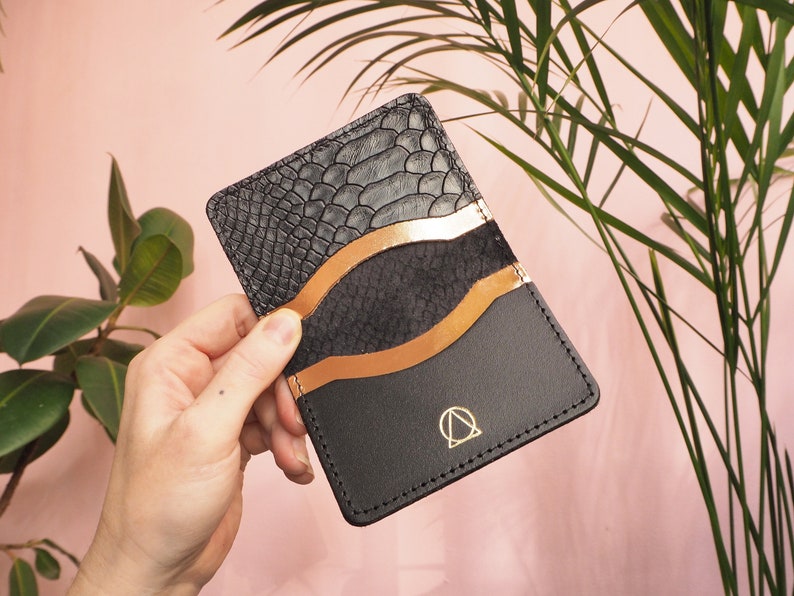 K&ouml;nnte beinhalten: Ein schwarzer Lederkartenhalter mit Schlangenhautmuster auf der vorderen Klappe. Der Kartenhalter hat einen kupferfarbenen Akzent und ein goldenes Logo mit einem Kreis und einem Dreieck.