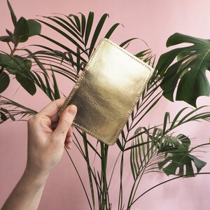K&ouml;nnte beinhalten: Ein goldfarbener Lederkartenhalter mit gen&auml;htem Rand, in einer Hand vor einem rosa Hintergrund mit gr&uuml;nen Pflanzen gehalten.