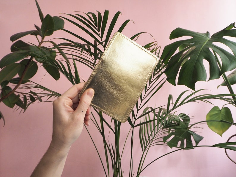K&ouml;nnte beinhalten: Ein goldfarbener Lederkartenhalter mit gen&auml;htem Rand, in einer Hand vor einem rosa Hintergrund mit gr&uuml;nen Pflanzen gehalten.