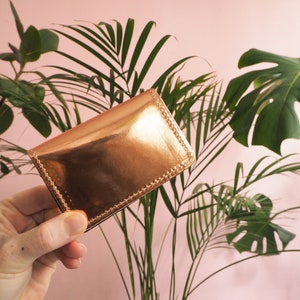 K&ouml;nnte beinhalten: Ein Kartenetui aus metallisch goldenem Leder mit einem gen&auml;hten Rand. Das Kartenetui wird in einer Hand gehalten.