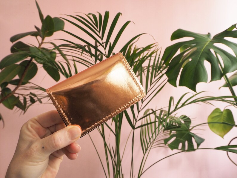 K&ouml;nnte beinhalten: Ein Kartenetui aus metallisch goldenem Leder mit einem gen&auml;hten Rand. Das Kartenetui wird in einer Hand gehalten.