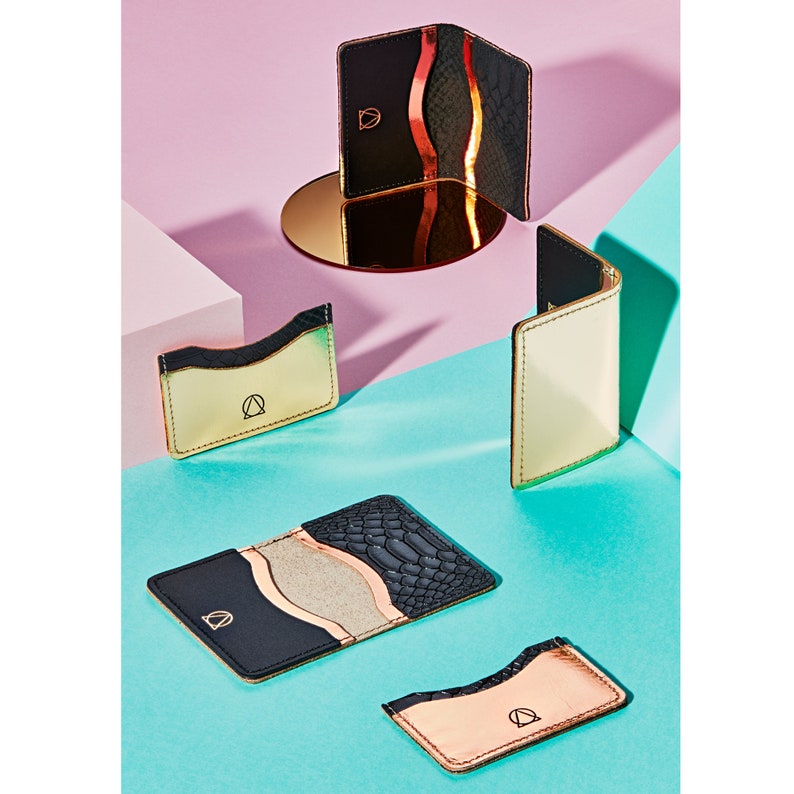 K&ouml;nnte beinhalten: Vier verschiedene Kartenhalter in verschiedenen Farben und Materialien. Die Kartenhalter sind aus Leder und haben ein goldenes Logo auf der Vorderseite. Die Kartenhalter sind auf einem rosa und blauen Hintergrund angeordnet.