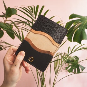 K&ouml;nnte beinhalten: Ein schwarzer Lederkartenhalter mit Schlangenhautmuster auf der oberen Klappe. Der Kartenhalter hat einen goldenen Akzent und ein goldenes Logo auf der Vorderseite.