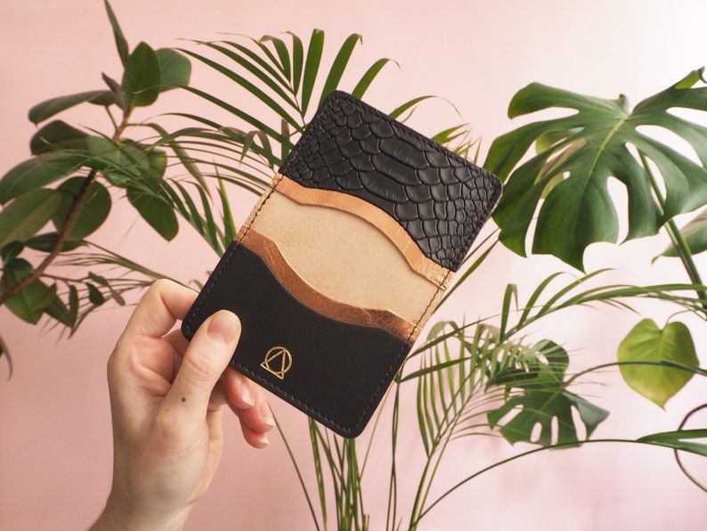 K&ouml;nnte beinhalten: Ein schwarzer Lederkartenhalter mit Schlangenhautmuster auf der oberen Klappe. Der Kartenhalter hat einen goldenen Akzent und ein goldenes Logo auf der Vorderseite.