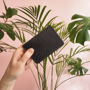 K&ouml;nnte beinhalten: Eine schwarze Lederb&ouml;rse mit strukturierter Oberfl&auml;che. Die B&ouml;rse wird in einer Hand gehalten.