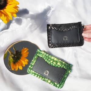 Może przedstawiać: Dwa małe czarne skórzane portfele z zamkiem błyskawicznym. Jeden portfel ma wzór wężowej skóry w kolorze zielonym, a drugi w kolorze czarnym. Oba portfele mają złote logo z przodu.