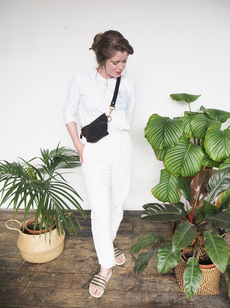 Op de afbeelding: Een vrouw draagt een wit overhemd met lange mouwen, een witte broek en zwart-wit gestreepte sandalen. Ze draagt een zwart leren heuptasje met een gouden ketting. De vrouw staat voor een witte muur met twee planten in potten.