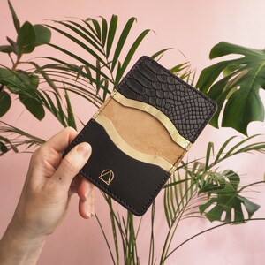 K&ouml;nnte beinhalten: Eine schwarze Ledergeldb&ouml;rse mit einem goldenen geometrischen Design und einem Schlangenhautmuster auf der Vorderseite. Die Geldb&ouml;rse hat mehrere Kartenf&auml;cher und ein Fach f&uuml;r Bargeld.