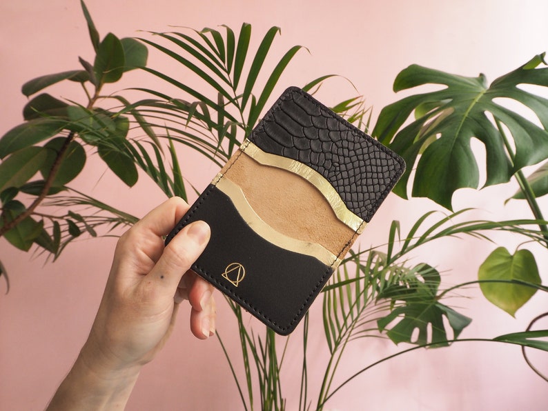 K&ouml;nnte beinhalten: Eine schwarze Ledergeldb&ouml;rse mit einem goldenen geometrischen Design und einem Schlangenhautmuster auf der Vorderseite. Die Geldb&ouml;rse hat mehrere Kartenf&auml;cher und ein Fach f&uuml;r Bargeld.