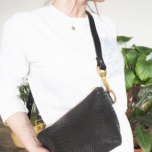 May include: A black leather crossbody bag with a snakeskin pattern and gold hardware. The bag is worn by a person with a white shirt and a silver necklace.