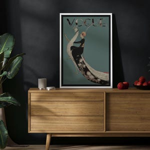 May include: A framed Vogue magazine cover featuring an illustration of a woman in a black dress and a feathered headdress, set against a teal background. The word "VOGUE" is at the top. The artwork is displayed on a wooden cabinet.