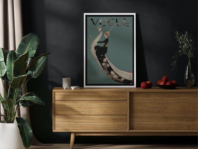 May include: A framed Vogue magazine cover featuring an illustration of a woman in a black dress and a feathered headdress, set against a teal background. The word "VOGUE" is at the top. The artwork is displayed on a wooden cabinet.