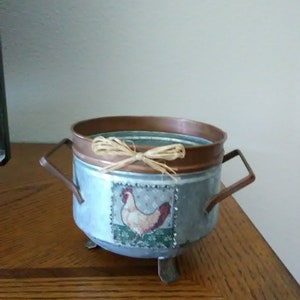 May include: A galvanized metal container with a copper rim and handles. The container features a decorative image of a hen and is tied with raffia. The container is on three small metal feet. Rustic farmhouse decor.