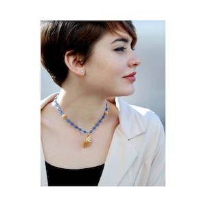 May include: A woman with short brown hair is wearing a necklace with a gold seashell pendant and blue beads. She is wearing a white blazer and a black top.