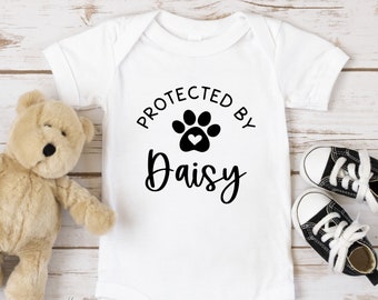 May include: White baby onesie with black text that reads "Protected by" with a black paw print with a white heart in the center and the name "Daisy" written in cursive below the paw print.