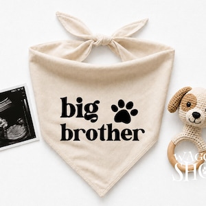May include: A beige fabric pet bandana featuring the black text big brother and a paw print icon, accompanied by a black and white ultrasound scan and a small crocheted dog rattle with a light brown and cream head and a wooden ring handle.