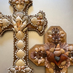 May include: Two wooden crosses decorated with seashells. The larger cross has a more intricate design with smaller shells, while the smaller cross has larger shells.